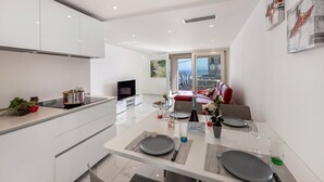 A modern kitchen with white cabinets, a dining area with a table set for two, and a living area with a red sofa and a TV. - Red View Apartment in Ronco Sopra Ascona Ascona (Ronco sopra Ascona)