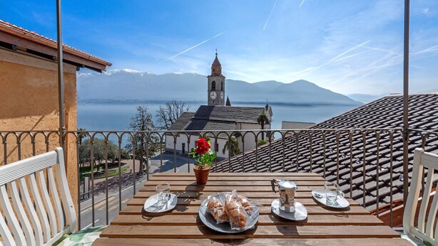 A terrace with a table set for two, overlooking a mountainous landscape and a body of water. - Red View Apartment in Ronco Sopra Ascona Ascona (Ronco sopra Ascona)