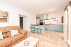 A modern living room with a sofa and a coffee table, a kitchen with a central island and bar stools, and a herringbone-patterned floor.