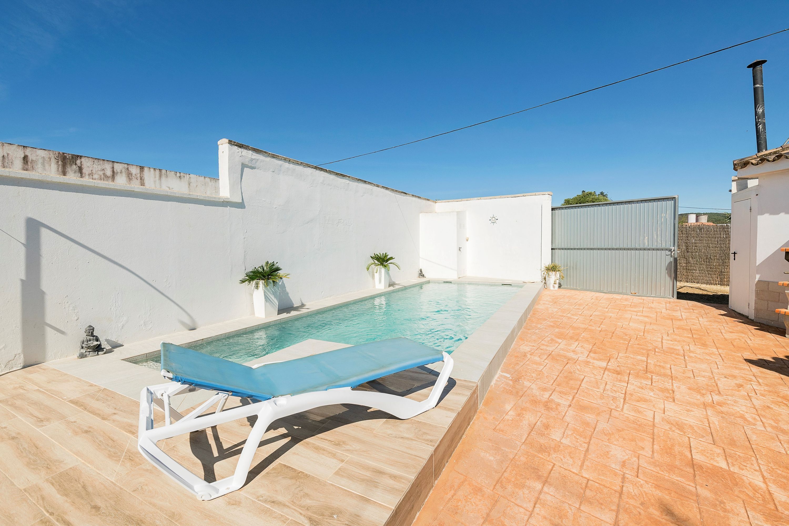 A swimming pool with a blue lounge chair, a white wall, and a tiled patio area.