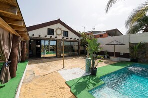 A pool area with a brick patio, a wooden pergola, and a house with a stone facade and a large window. - Casa Amelie in Conil de la Frontera (Conil de la Frontera)