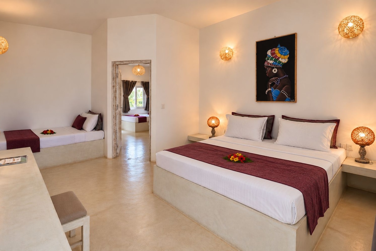 A hotel room with a large bed, two smaller beds, a desk, and a chair. There are decorative wall sconces and a painting on the wall.