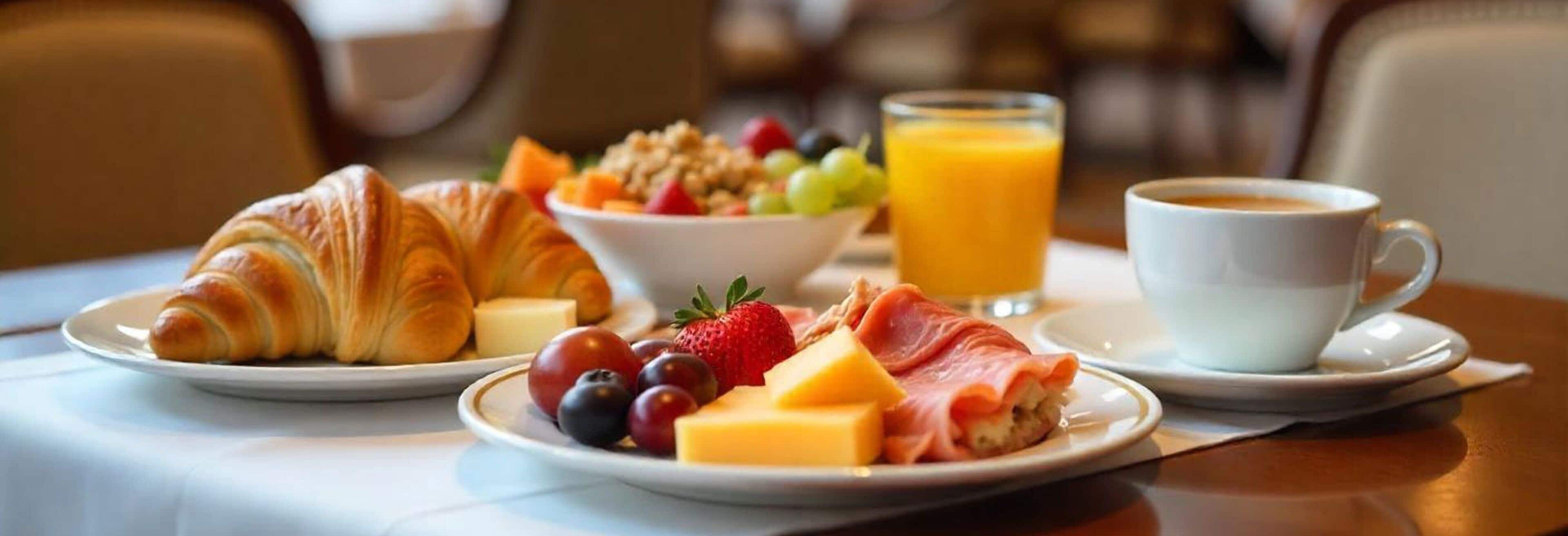 A breakfast setting with croissants, cheese, ham, fruits, a bowl of cereal, orange juice, and a cup of coffee on a table.