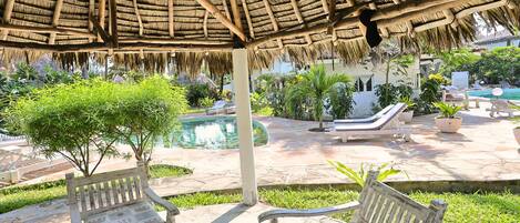 A covered outdoor area with a pool, lounge chairs, and a thatched roof structure.