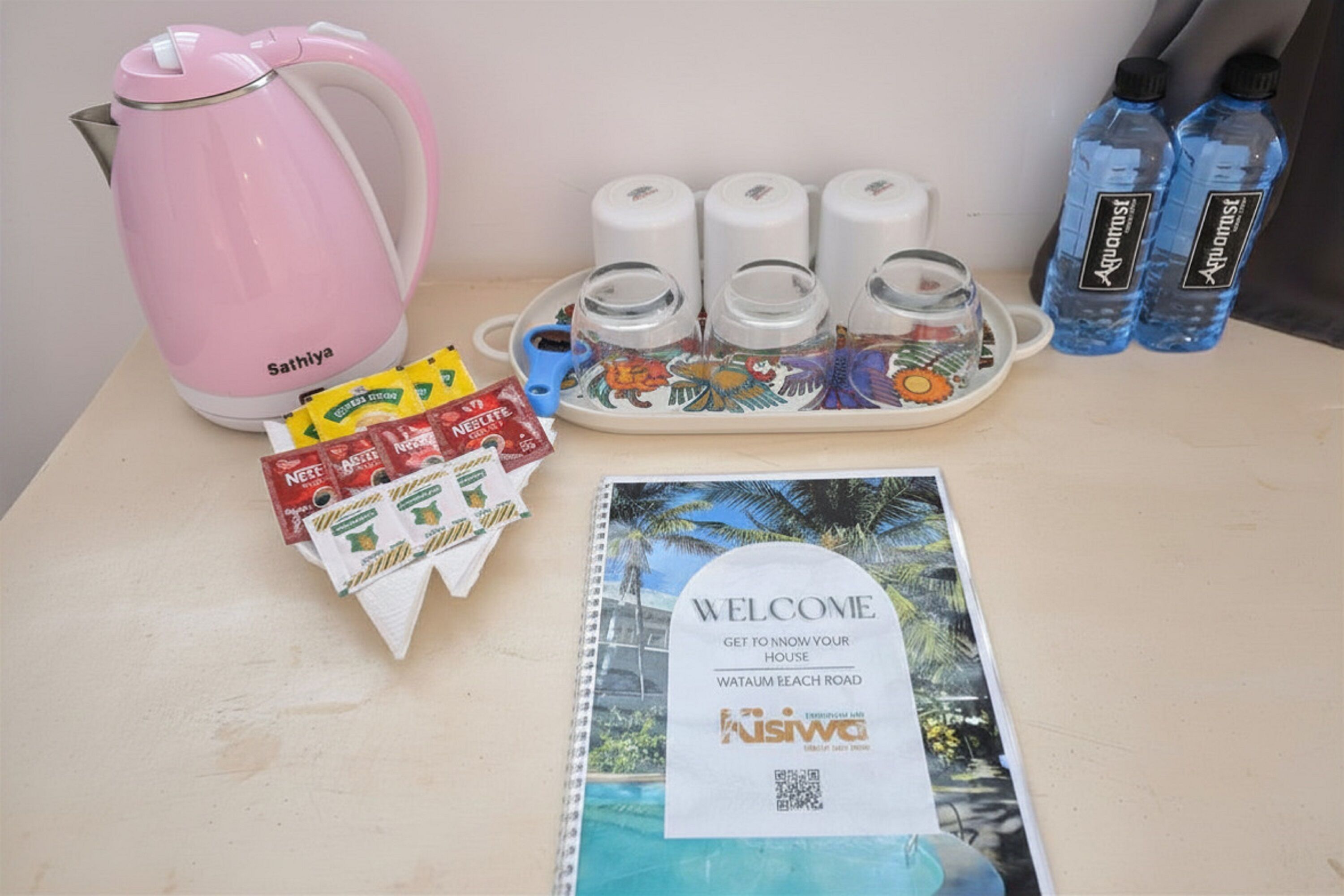 A table with a pink kettle, water bottles, a tray with cups, a welcome brochure, and some coffee packets.