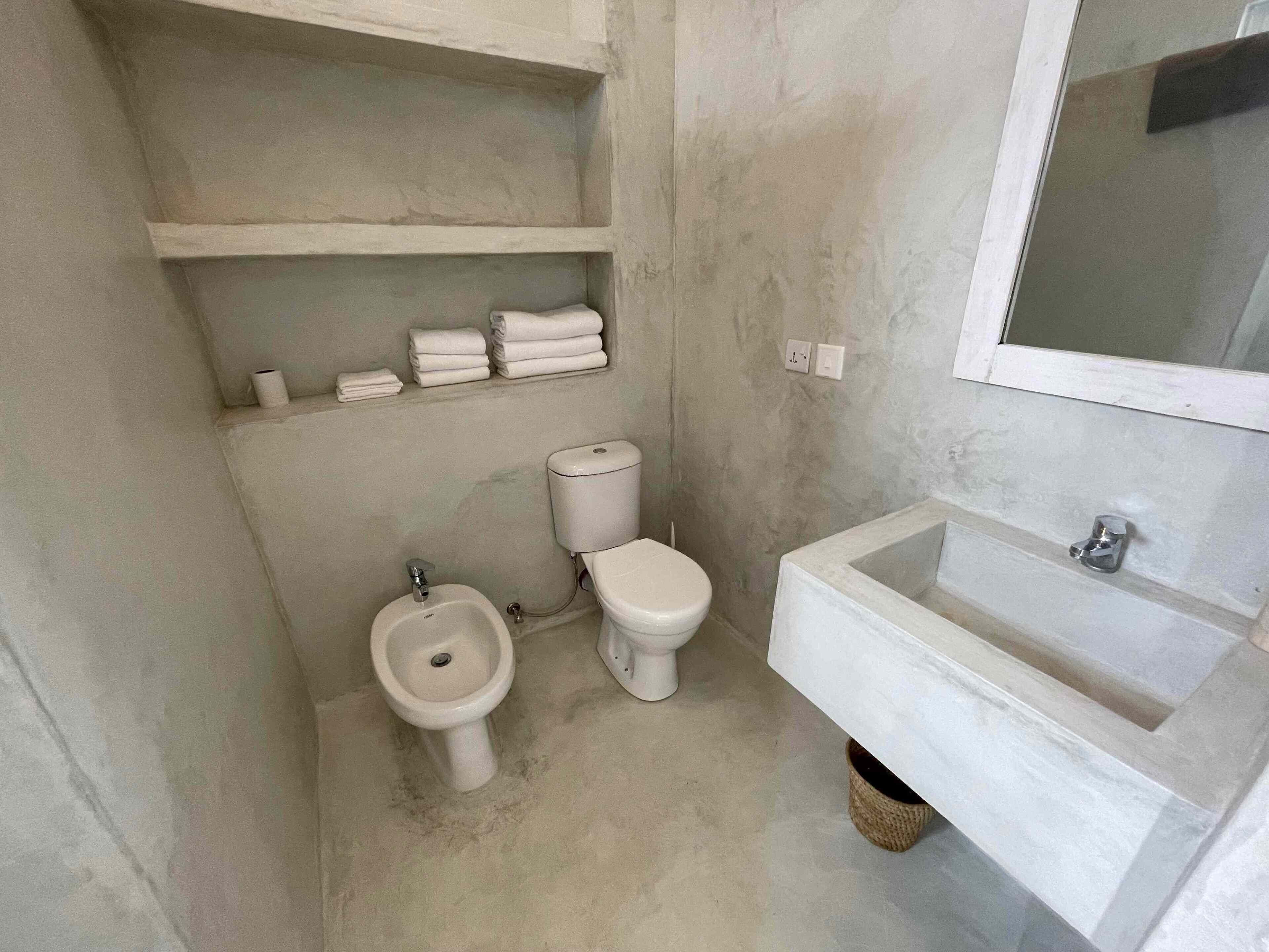 Een kleine badkamer met toilet, bidet en wastafel. Er staan planken met opgevouwen handdoeken en een klein afvalbakje.