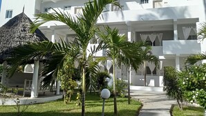 Un edificio bianco con numerosi balconi, una struttura con tetto di paglia e un giardino curato con palme.