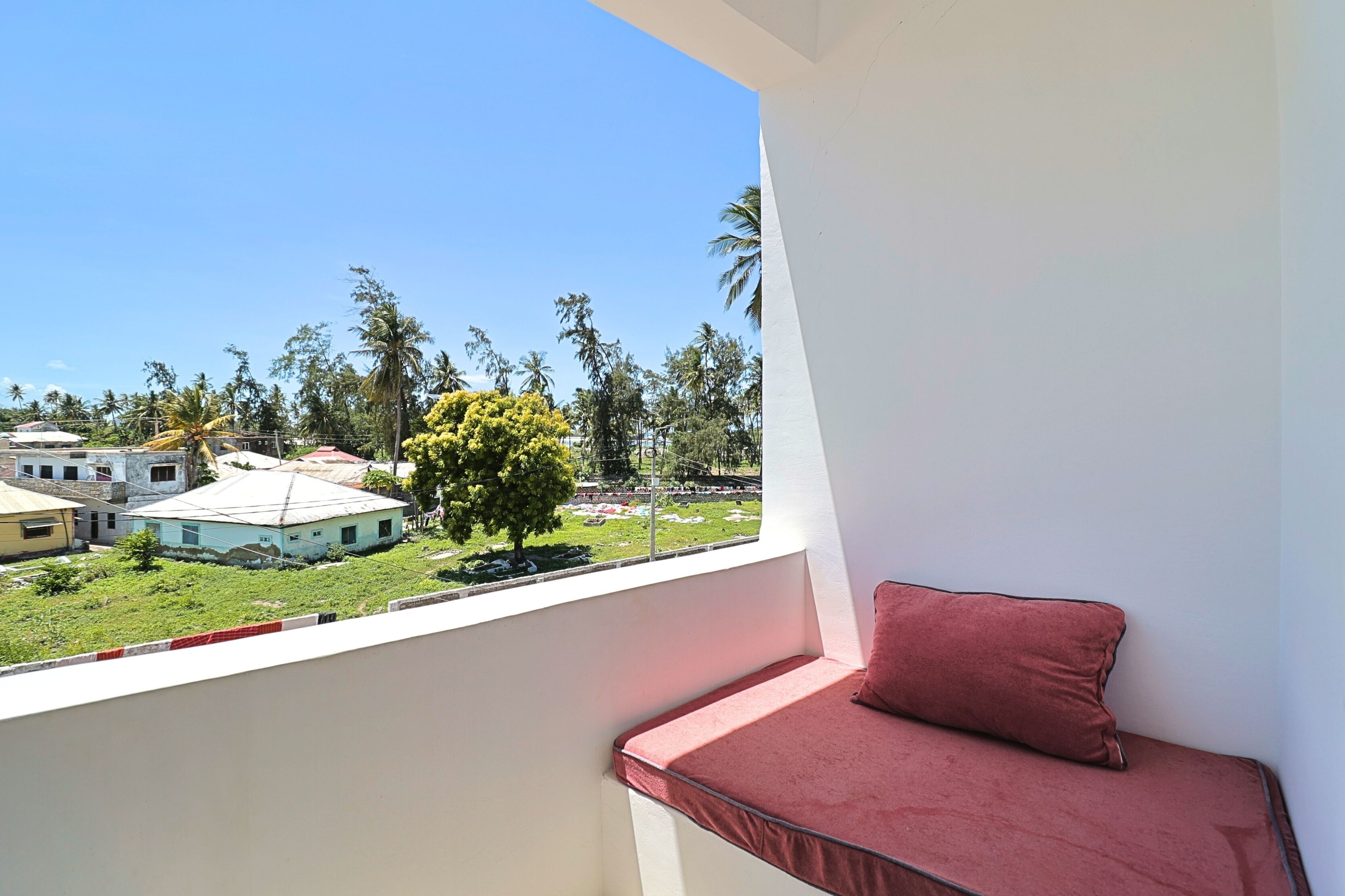 A balcony with a red cushion, a view of a building with a blue roof, and palm trees.