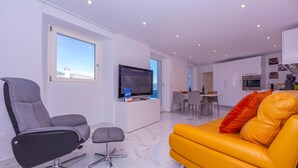 Ein modernes Wohnzimmer mit großem Fernseher, einem gelben Sofa, einem grauen Bürostuhl und einem Essbereich im Hintergrund.