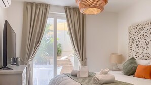 A hotel room with a bed, a desk with a flat-screen TV, a lamp, and a view of the outdoors. - Modern Beautifull Pool And Sea Apt Qawra (St. Paul's Bay)