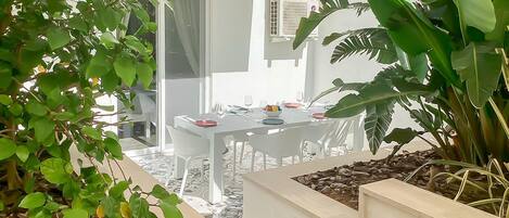 Un comedor con una mesa preparada para dos personas, rodeada de sillas blancas, y vista a un edificio en el fondo.