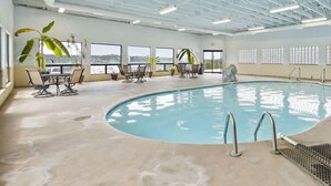 Une piscine intérieure offrant une vue dégagée sur la ville grâce à de grandes fenêtres, entourée de chaises longues et de plantes en pot.