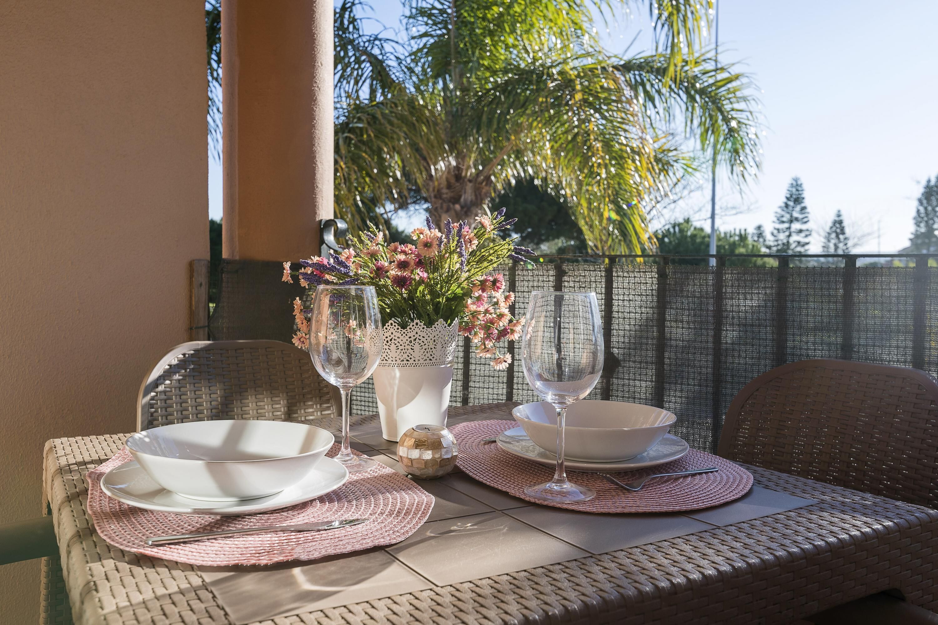 A table set with white bowls, wine glasses, and a vase of flowers on a wicker table outdoors.