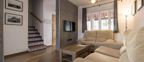 A living room with a sofa, a coffee table, a TV mounted on the wall, and a staircase with patterned steps.