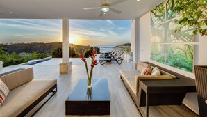 Una sala moderna con vistas a la puesta de sol sobre el océano, un ventilador de techo y un ventanal con un estampado de motivos naturales.