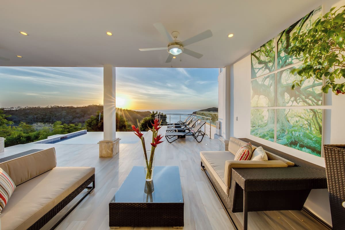 Un salon moderne avec vue sur le coucher de soleil sur l’océan, doté d’un ventilateur de plafond et d’une grande fenêtre ornée d’un motif inspiré de la nature.