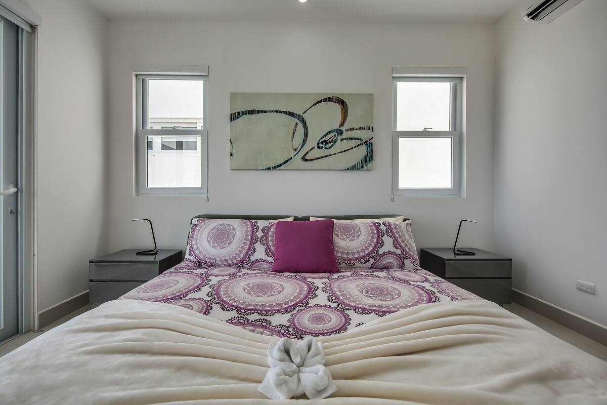 Une chambre à coucher comprenant un lit, deux tables de chevet, une commode, une fenêtre et un tableau abstrait accroché au mur.