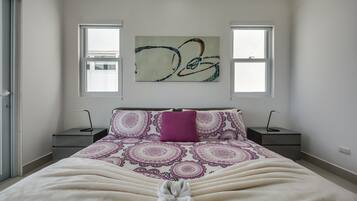 Un dormitorio con cama, dos mesitas de noche, una mesilla y una ventana, además de un cuadro abstracto colgado en la pared.
