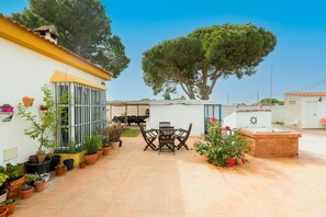 Un patio con una mesa y sillas, rodeado de macetas con plantas y un árbol grande.