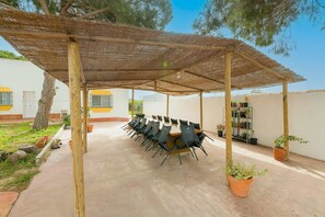 A covered outdoor seating area with a long table and chairs, surrounded by potted plants and a white building with yellow accents. - El Coto in Chiclana de la Frontera (Chiclana de la Frontera)