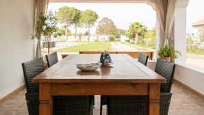 A wooden table with wicker chairs, a teapot, and a plate on a patio with a view of a garden and trees. - El Coto in Chiclana de la Frontera (Chiclana de la Frontera)