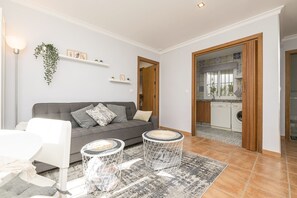 Un salon moderne avec un canapé gris, des fauteuils blancs et deux petites tables d’appoint rondes. Une porte en bois donne accès à un espace cuisine.