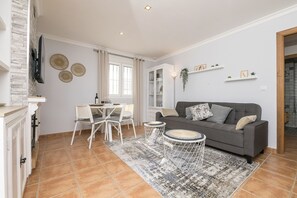 Un salon avec un canapé gris, une table à manger blanche et des chaises. On y trouve un téléviseur fixé au mur, une cheminée et une fenêtre avec des rideaux.