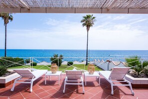 A terrace with white lounge chairs and tables, overlooking a clear blue sea and palm trees. - Villa Acqua Saracena by Wonderful Italy (Augusta)