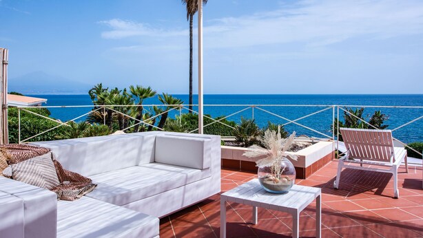 Een terras met een witte bank, een houten stoel en een tafel met een decoratieve schaal. Het terras heeft uitzicht op een heldere blauwe zee en is voorzien van een rieten dak.