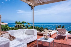 A terrace with a white sofa, a wooden chair, and a table with a decorative bowl. The terrace overlooks a clear blue sea and features a thatched roof. - Villa Acqua Saracena by Wonderful Italy (Augusta)