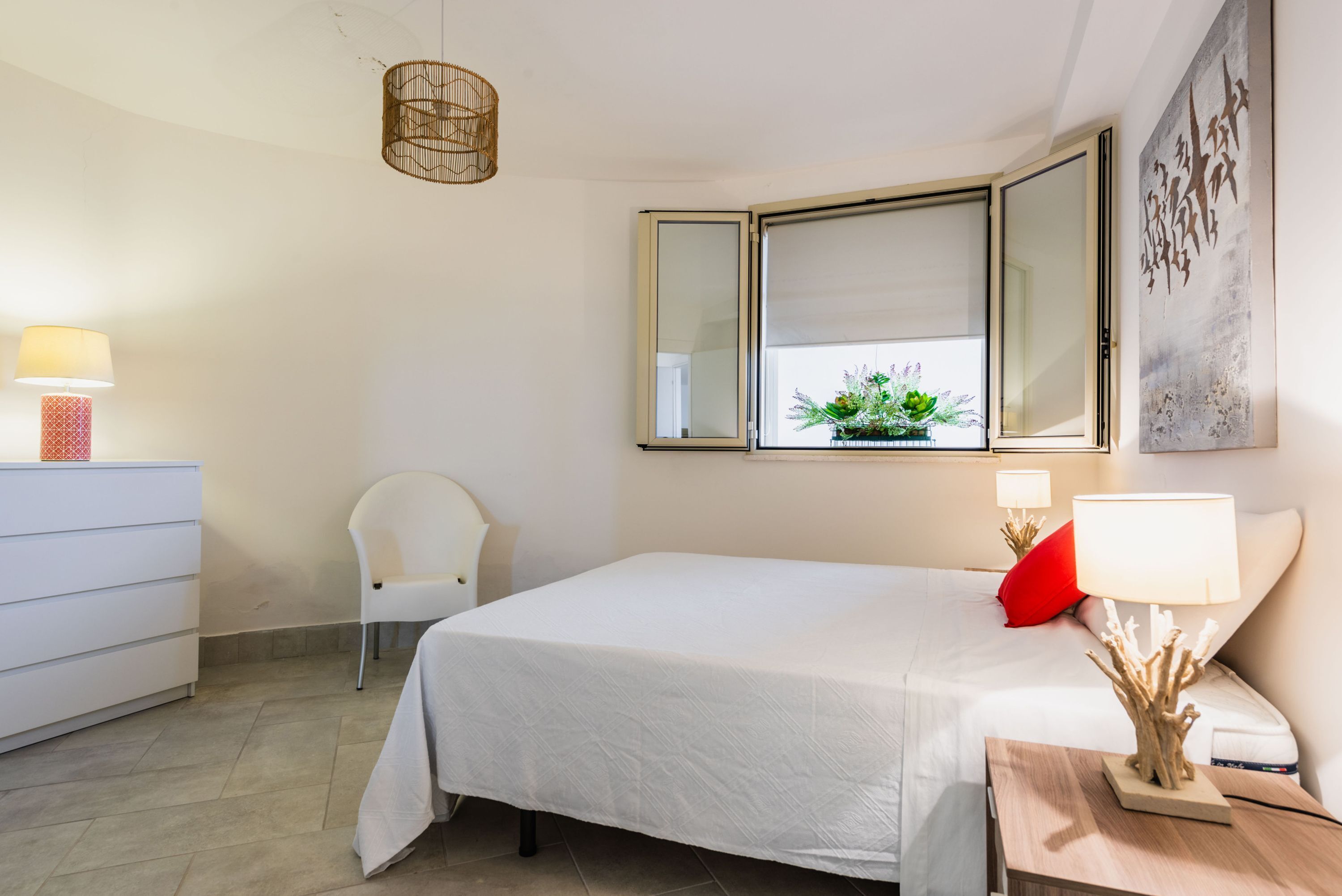 A bedroom with a bed, bedside lamps, a chair, a dresser, and a window with a view of greenery.