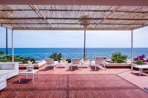 A covered outdoor seating area with a view of the sea, featuring white lounge chairs, a white sofa, and a small table with a decorative item. - Villa Acqua Saracena by Wonderful Italy (Augusta)
