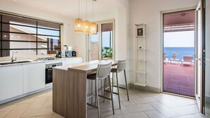 Een moderne keuken met een groot eiland, barstoelen en uitzicht op de zee door een open deur.
