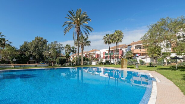 A swimming pool with a tiled edge, surrounded by palm trees and buildings with red and beige facades. - Oasis Playero in Chiclana de la Frontera (Chiclana de la Frontera)