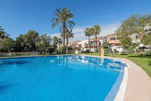 A swimming pool with a tiled edge, surrounded by palm trees and buildings with red and beige facades. - Oasis Playero in Chiclana de la Frontera (Chiclana de la Frontera)
