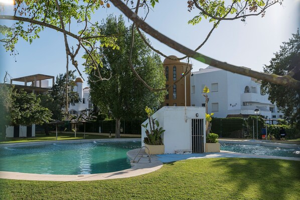 Une piscine entourée de pelouse et d’arbres bien entretenus, avec des bâtiments en arrière-plan.