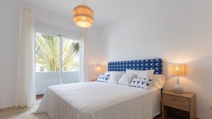 A bedroom with a large bed, two bedside lamps, a nightstand, and a view of palm trees through the window. - Saladillo in Estepona (Estepona)