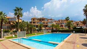 A rectangular swimming pool with a tiled deck, surrounded by palm trees and a row of multi-story buildings with balconies. - Perseo in Torrevieja (Torrevieja)