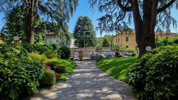 A stone pathway flanked by lush greenery and trees, leading to a gated entrance with parked cars. - Rovani 14 in Monza With 1 Bedrooms and 1 Bathrooms (Monza)