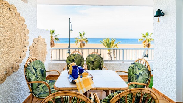 Un coin repas avec vue sur la plage, des chaises en osier, une table recouverte d’une nappe à carreaux et un élément décoratif mural.