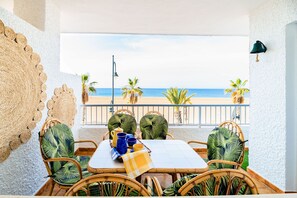 Un coin repas avec vue sur la plage, des chaises en osier, une table recouverte d’une nappe à carreaux et un élément décoratif mural.
