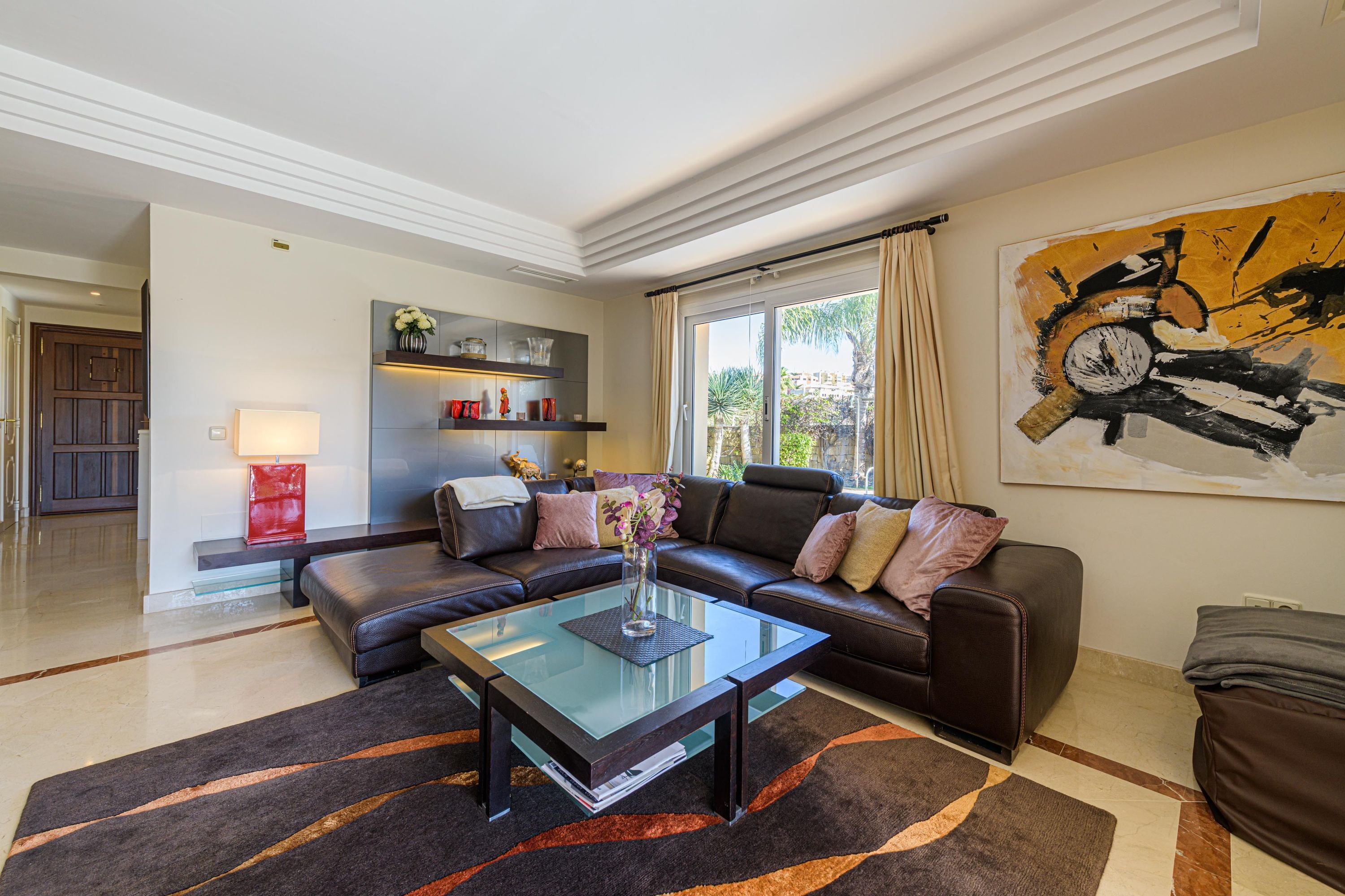 A modern living room with a glass coffee table, leather sofa, and a large abstract painting.