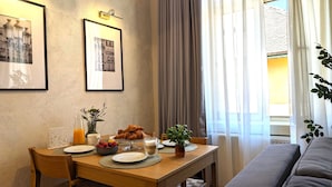 Ein Essbereich mit einem Holztisch, gedeckt für das Frühstück, inklusive Croissants, Obst und einem Kaffeedrink. An der Wand befinden sich gerahmte Bilder, eine Topfpflanze und ein Fenster mit Vorhängen.
