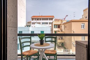 Un balcón con una pequeña mesa y sillas, una planta en maceta y vistas a edificios al fondo.