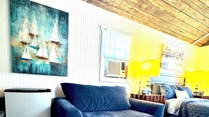 A room with a blue sofa, a white cabinet, a painting of sailboats, a window with blinds, and a bed with a blue quilt.