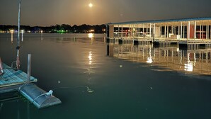A calm waterfront with a dock and a building with large windows reflecting the moonlight.