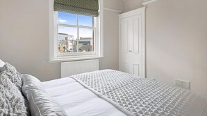 A bedroom with a bed, a window with a patterned valance, a white door, and a radiator. - Serene 2BR - 3 Mins to Fulham Broadway (London)