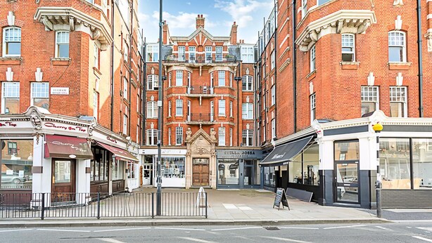 A street view of a red brick building with multiple storefronts, including a visible restaurant and a shop with a sign that reads 'Stella Dossa.' - Serene 2BR - 3 Mins to Fulham Broadway (London)
