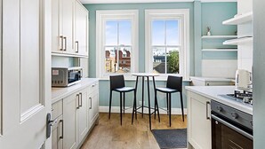 A modern kitchen with white cabinets, a microwave, a stove, and two bar stools. - Serene 2BR - 3 Mins to Fulham Broadway (London)