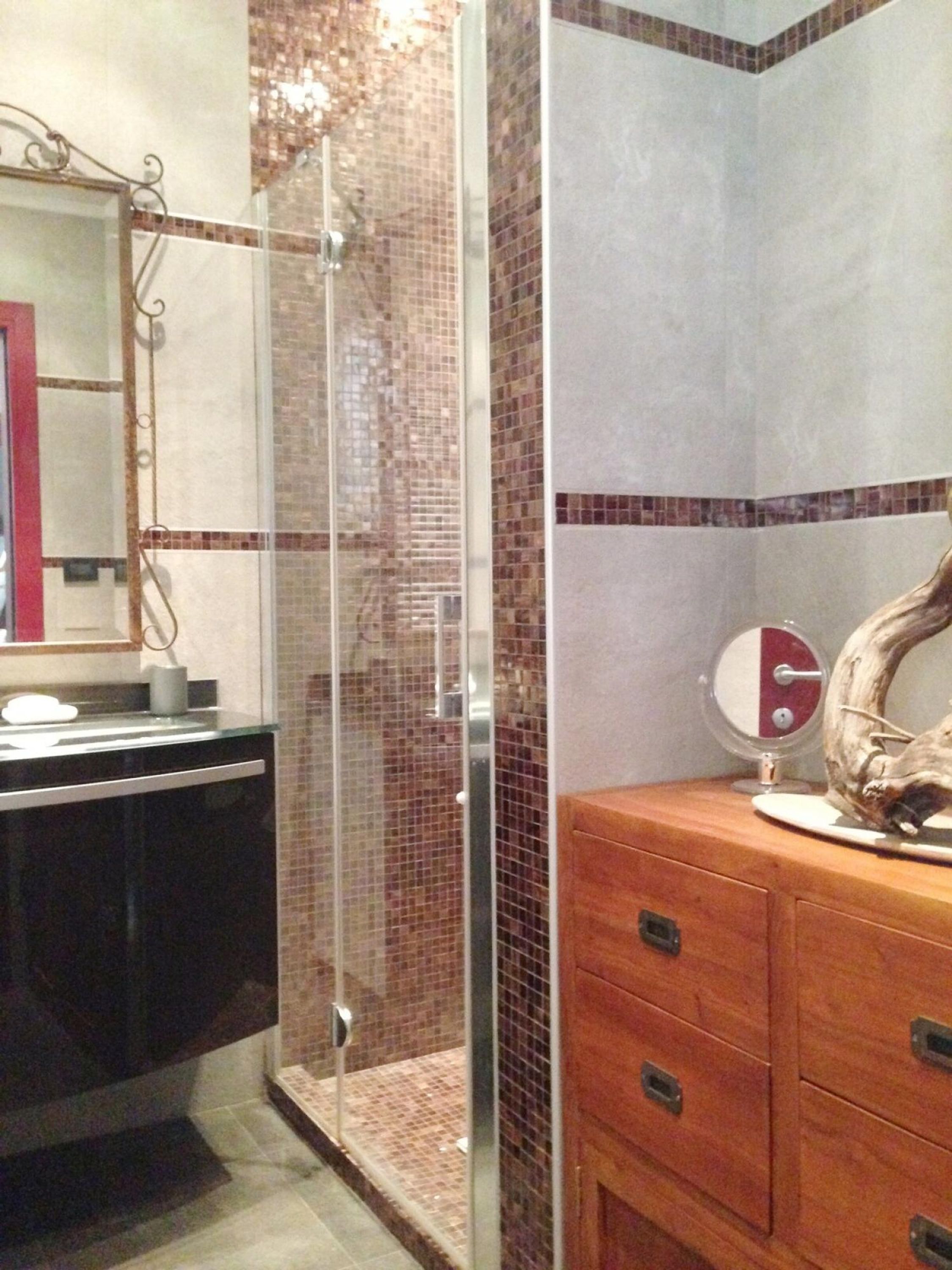 A bathroom with a glass shower enclosure, a wooden vanity with drawers, a round mirror, and a decorative piece on the vanity.